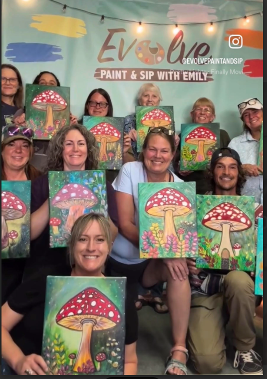 A full class holding up their mushroom paintings in front of the Evolve Paint & Sip banner