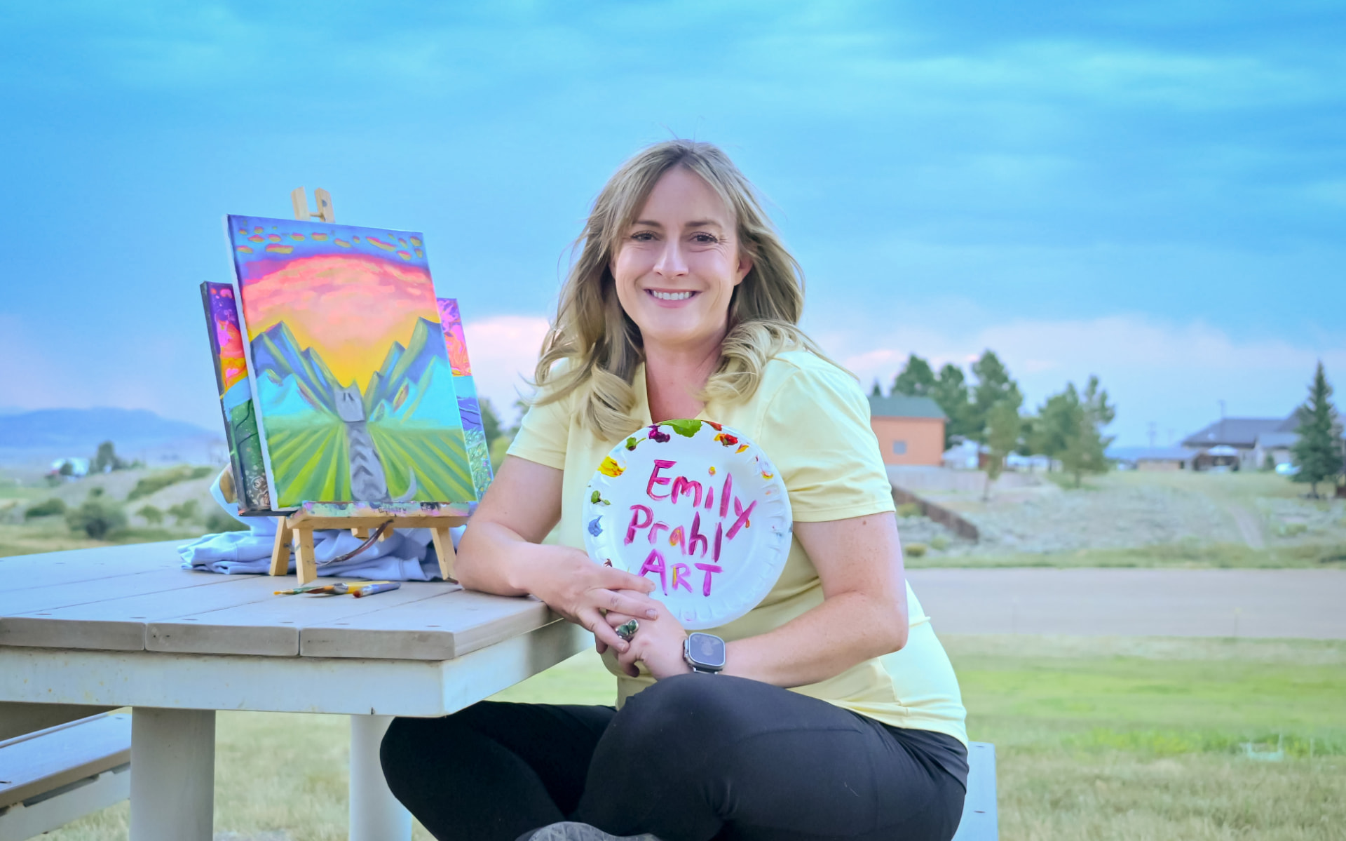 Emily Prahl outdoors with her painted palette and colorful paintings on an easel